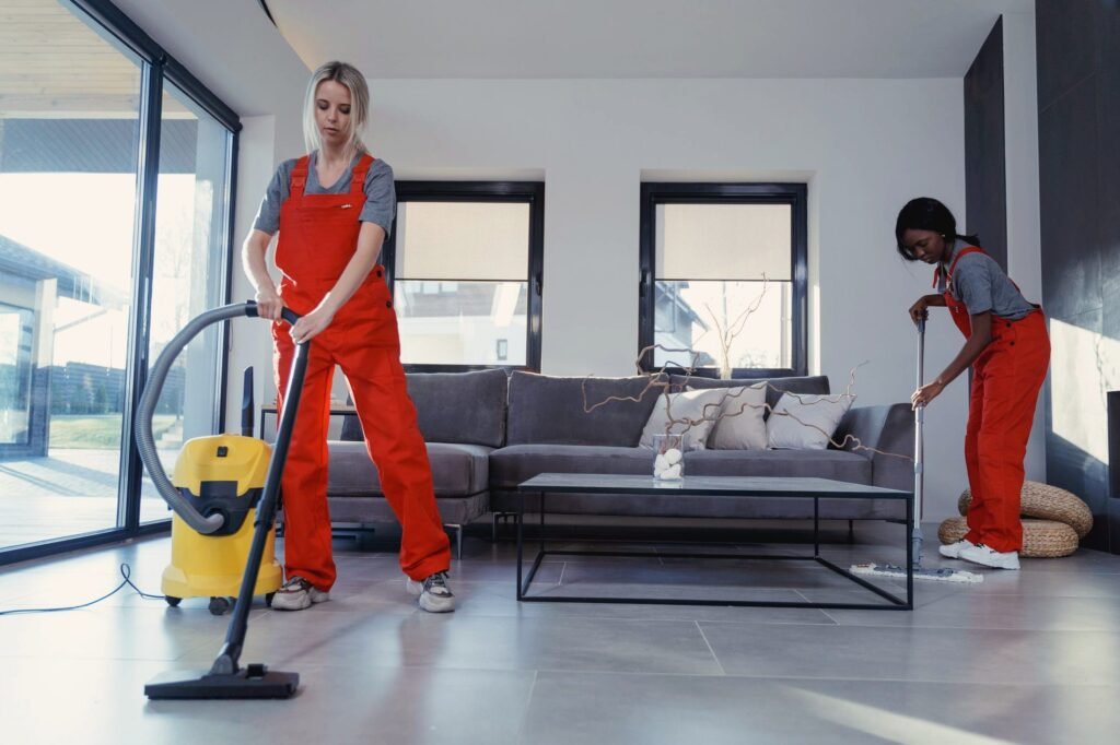 Zwei Reinigungskräfte in orangefarbener Arbeitskleidung saugen und wischen einen hellen Raum